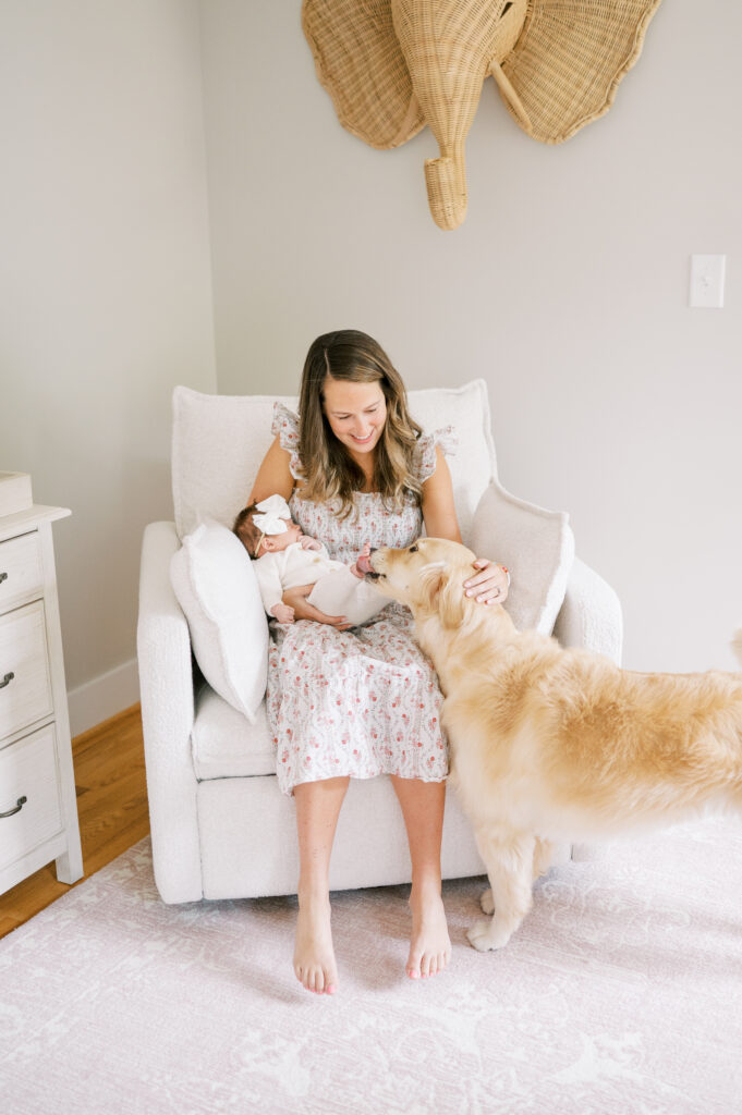 New mom holds baby girl in nursery rocking chair while golden retriever comes to sniff