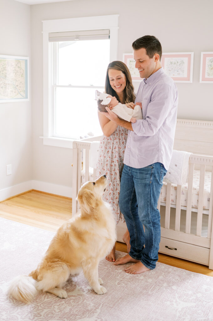 New parents hold their baby girl in front of crib in nursery while dog looks up at them