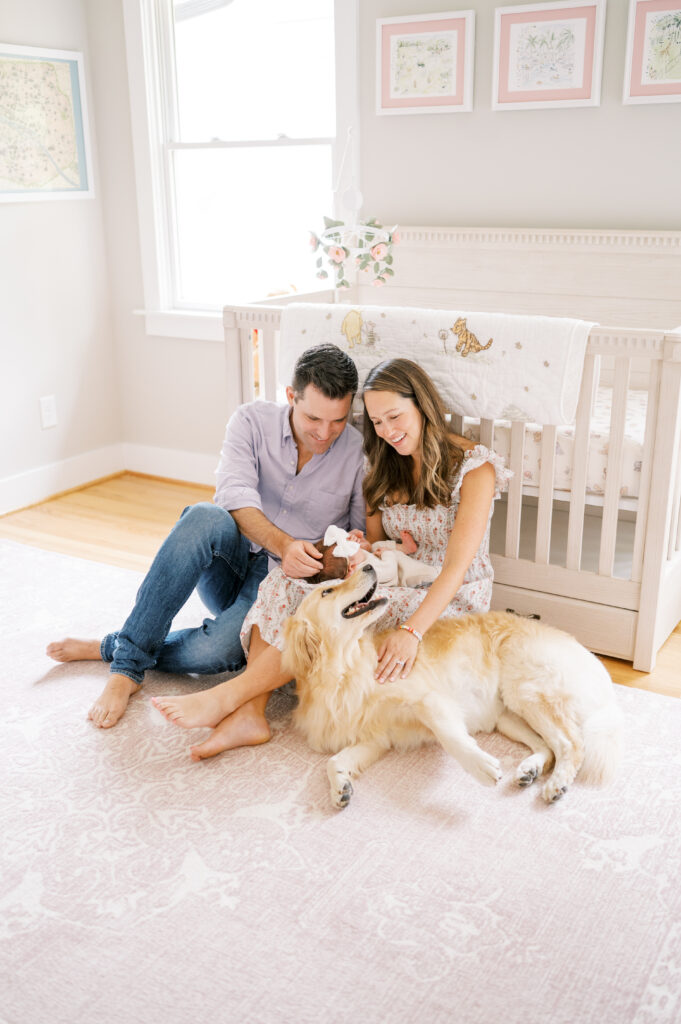 New parents sit in nursery with newborn baby girl and golden retriever during in-home photo session.