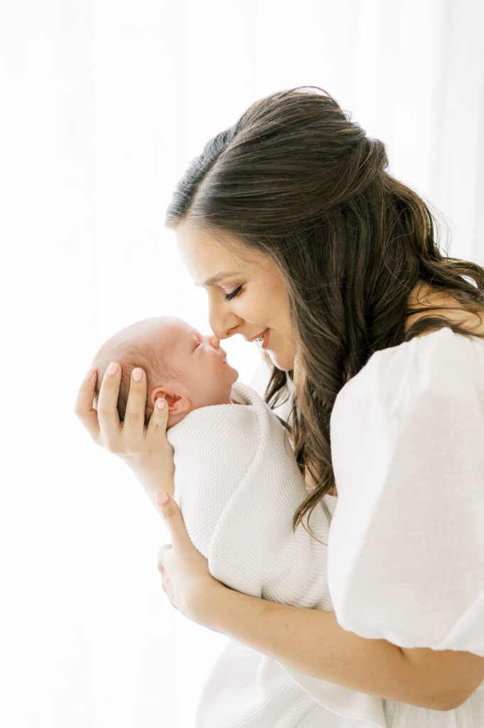 Mom in white dress holds swaddled newborn baby boy and touches her nose to his in Raleigh studio newborn session