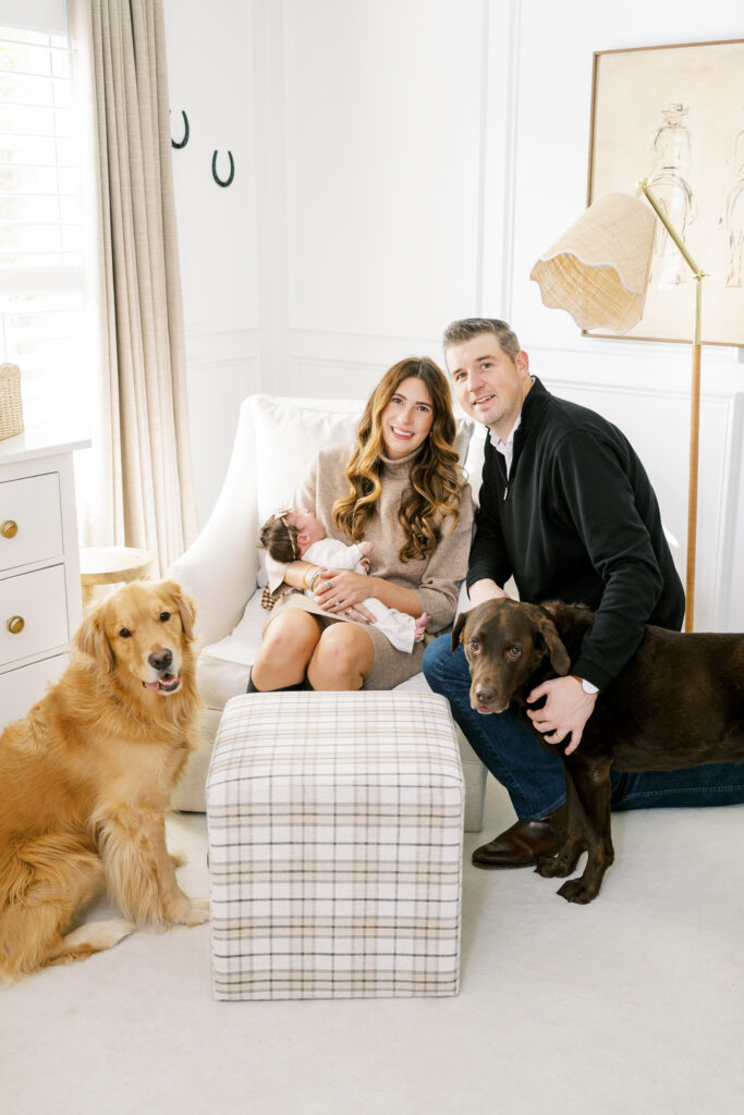 Parents hold baby girl and smile at camera with two dogs in nursery for newborn photo session