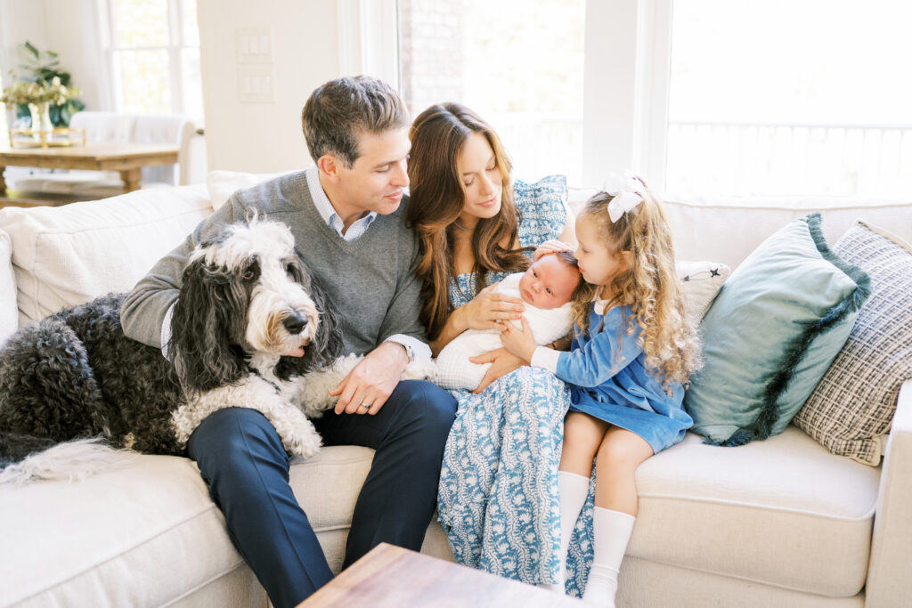 Family snuggles on couch with dog, swaddled newborn baby boy, and big sister cuddling him during Raleigh newborn session
