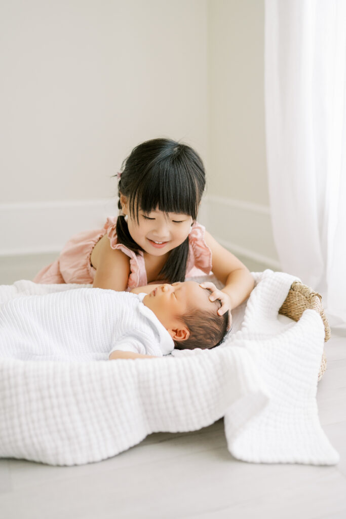 Big sister with pigtails and pink dress looks down at newborn baby brother laying in a basket during newborn photos with Worth Capturing Photography
