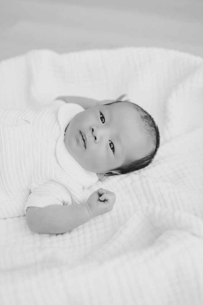 Black and white image of baby boy laying on a white blanket and looking at the camera during studio newborn session