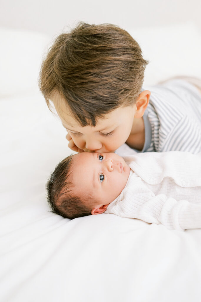 Big brother kisses baby sister laying on white bed during newborn photo session