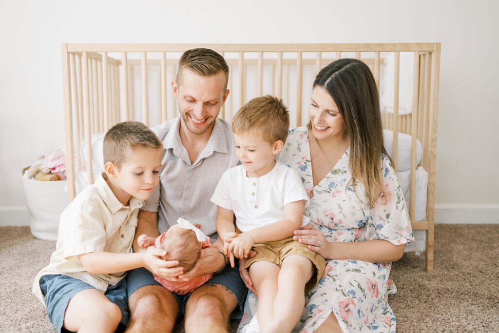 Family of five sits in front of crib smiling at newborn baby girl during in-home newborn photos