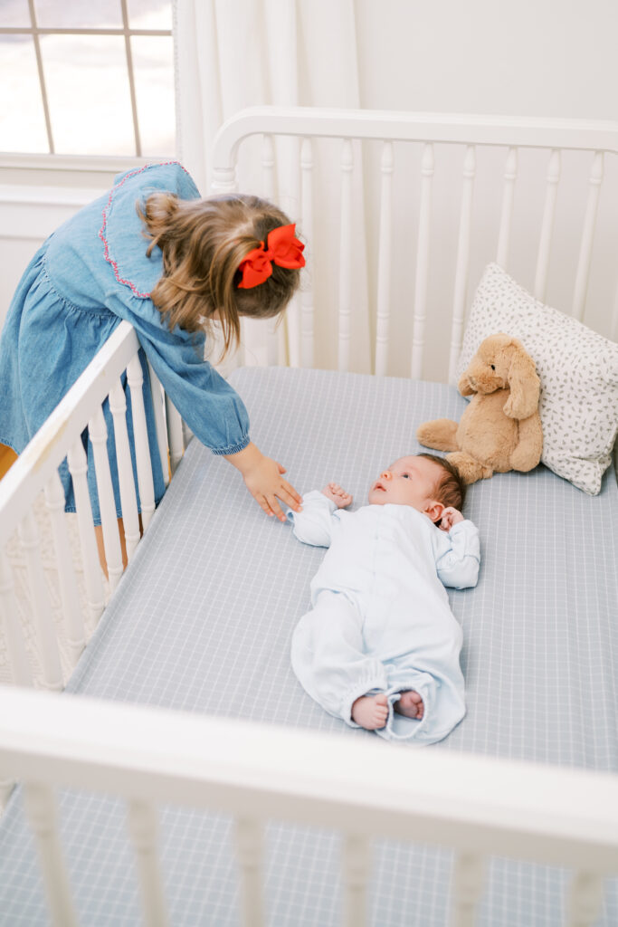 Big sister reaches into crib to hold hand of newborn baby brother during in-home newborn session with Worth Capturing Photography