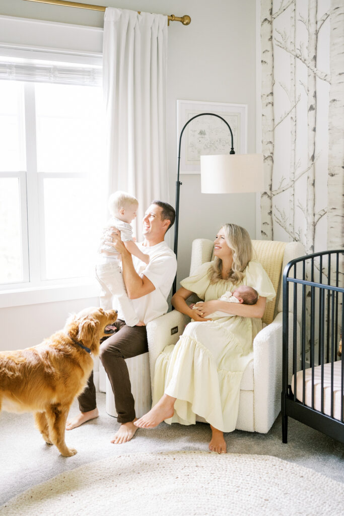 Mom holding newborn baby sits next to Dad lifting up toddler boy with golden retriever in nursery during newborn photos with Worth Capturing