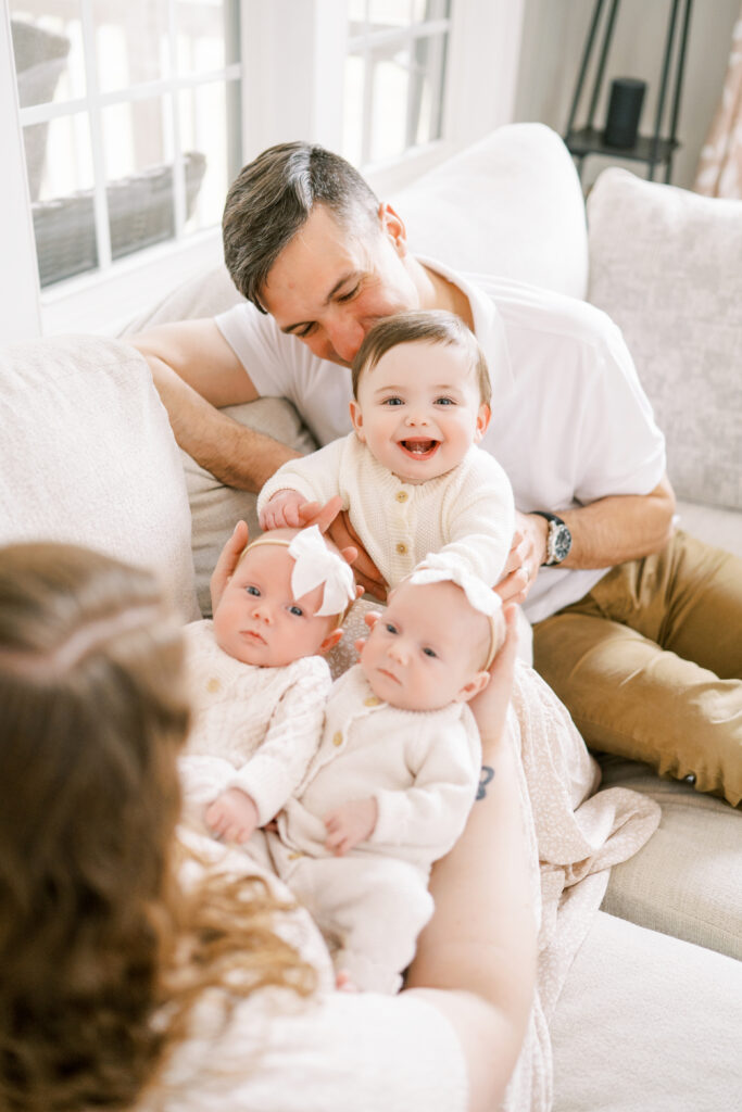 Baby girl smiles at camera while taking newborn photos with her new twin baby sisters and parents in Raleigh, NC
