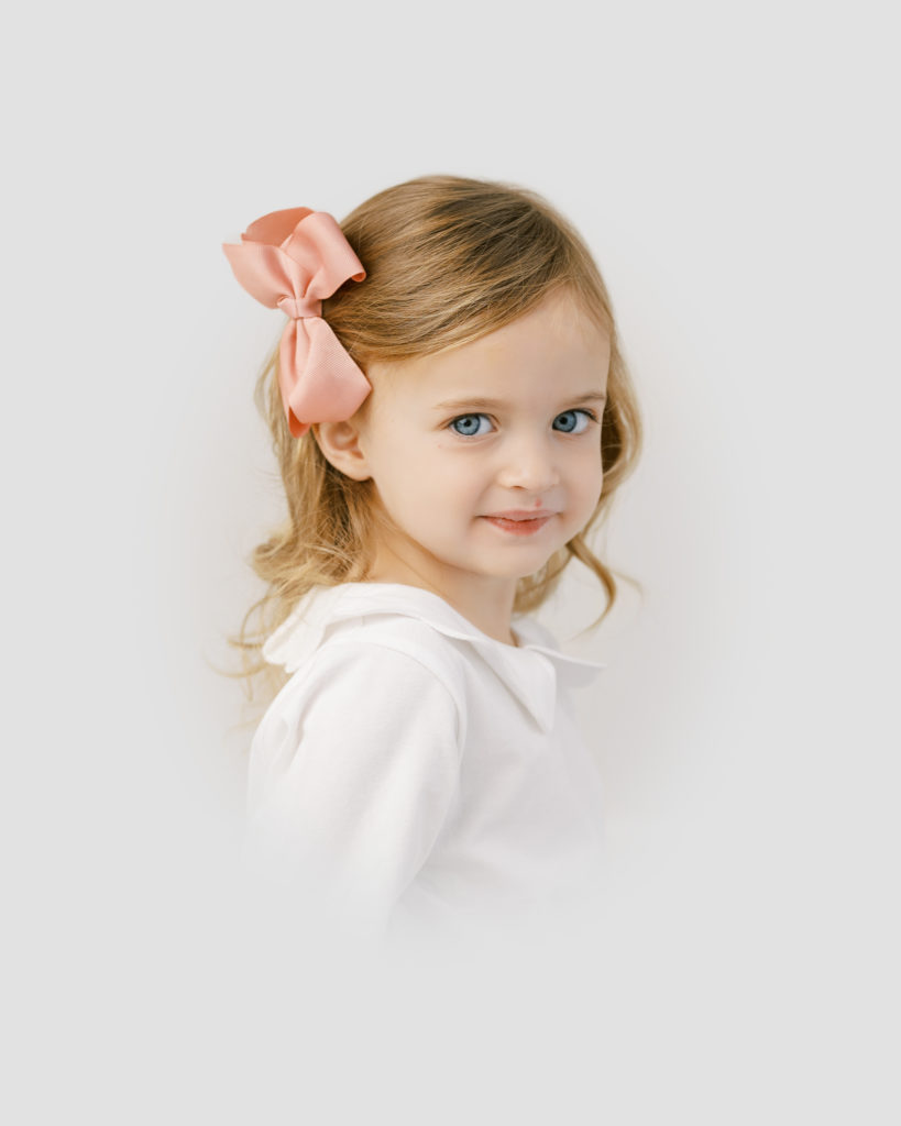 Heirloom studio portrait of little girl with brown curls, pink bow, and blue eyes by Worth Capturing photography in Raleigh, NC.