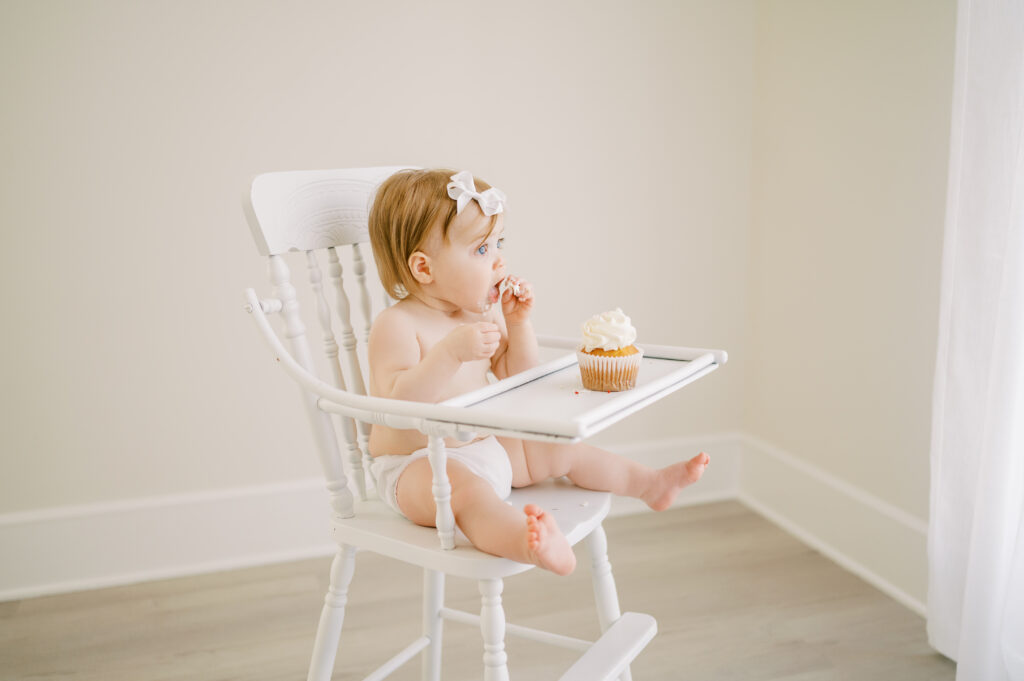 Baby girl in white diaper cover and bow sits in antique high chair eating a cupcake during first birthday photo session.