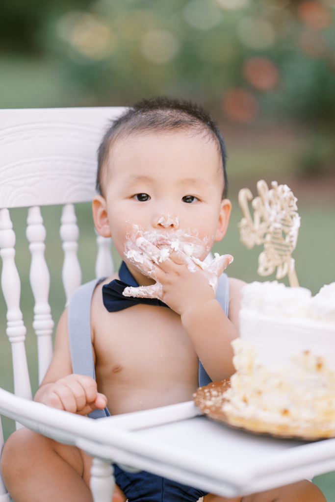 Baby eats cake frosting off of fingers while sitting in high chair during photography session in rose garden