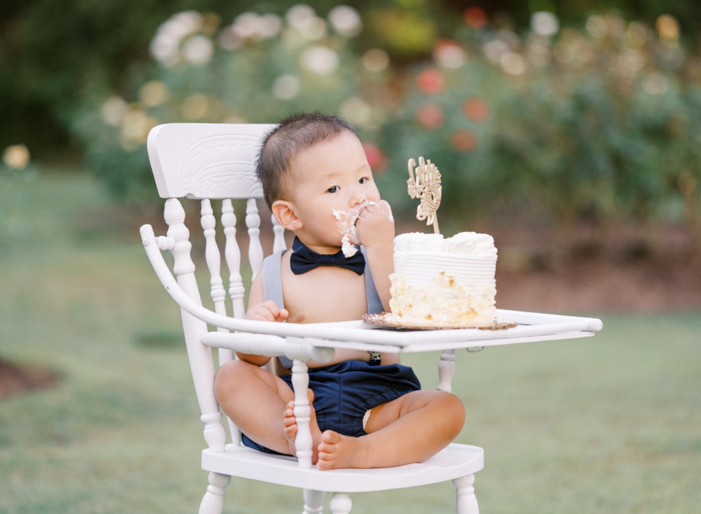 Baby boy in bowtie sits in rose garden in white high chair eating first birthday cake smash during photo session.