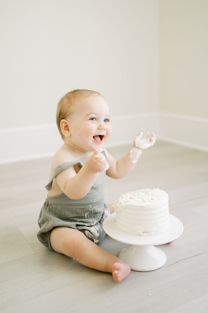 Baby boy sits with birthday cake and smiles during cake smash photography session at Worth Capturing Studio