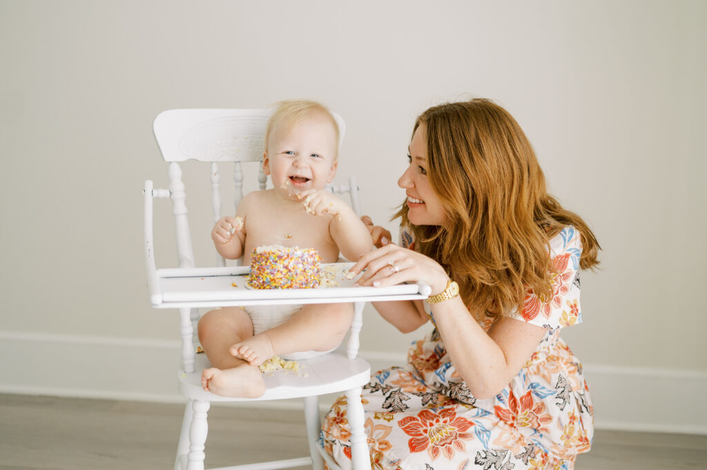 Baby sits in high chair with smashed birthday cake while mom stands beside and smiles at him.