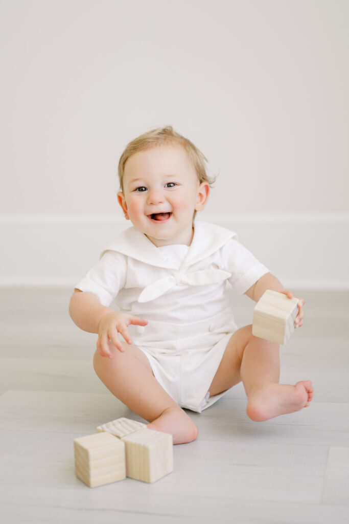 Baby boy in white sailor outfit plays with wooden blocks during his first birthday photos with Worth Capturing Photography.