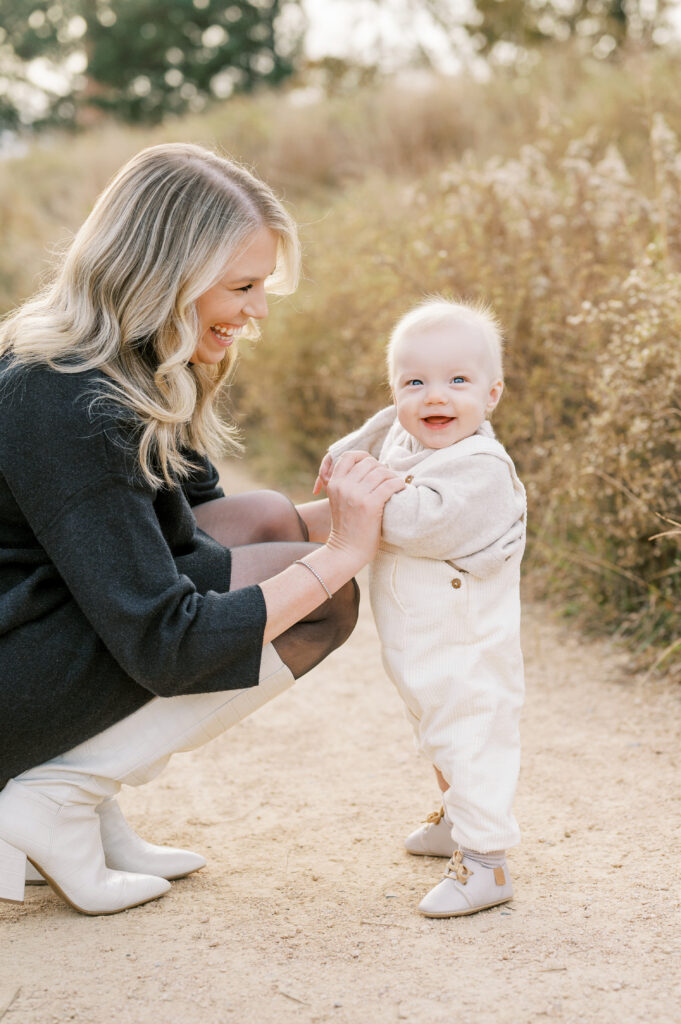 Baby boy stands on path and smiles with mom holding hands during family photos