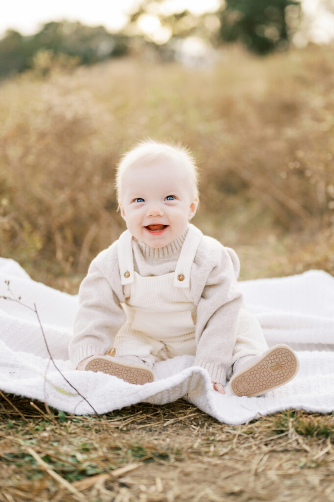 Baby boy sits in tall grass smiling during Fall family session in Raleigh
