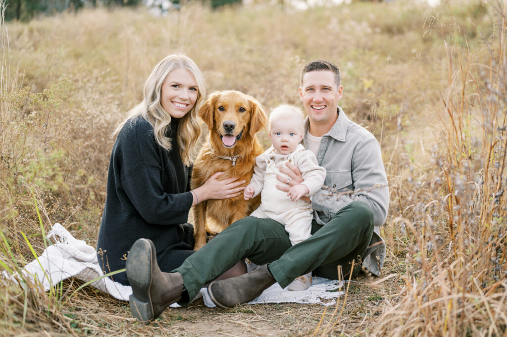 Mom and dad with baby boy and golden retriever look at camera during Fall family photo session in Raleigh.