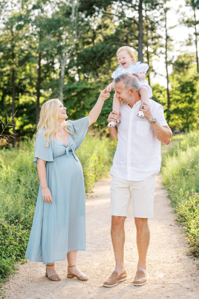 Pregnant mom in blue-green dress smiles at little girl sitting on dad's shoulders and holds her hand during family photos in Raleigh, North Carolina.
