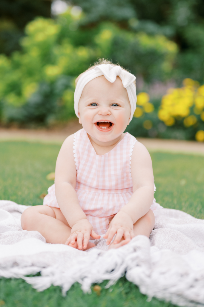 One year old baby girl wearing pink and white outfit and white headband sits on blanket and laughs during family photo session in Raleigh, NC