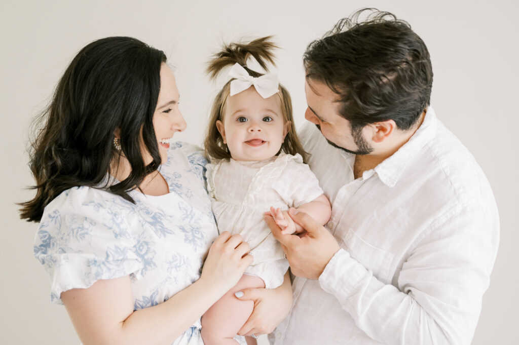 Mom and Dad hold 1 year old baby girl in white outfit during first birthday photo session at Worth Capturing Studio in Raleigh, North Carolina.