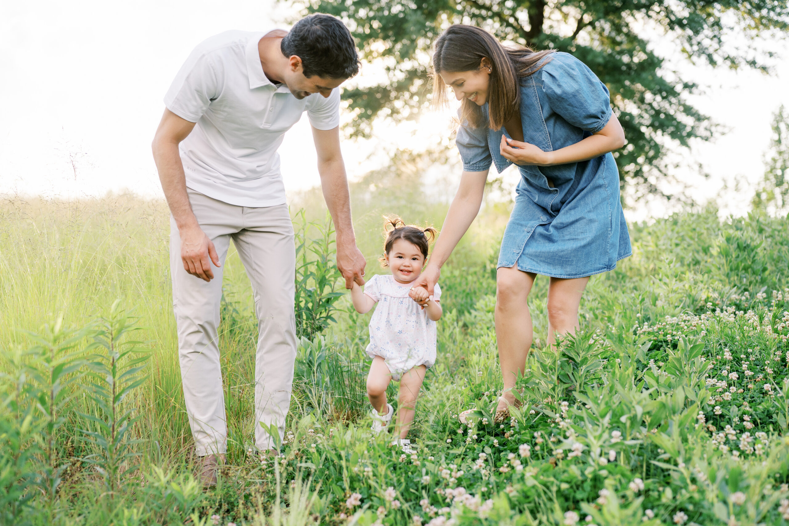 Mom in denim dress and Dad in white polo shirt hold hands of one year old little girl with pigtails walking through a green lush field with sun glowing behind them.
