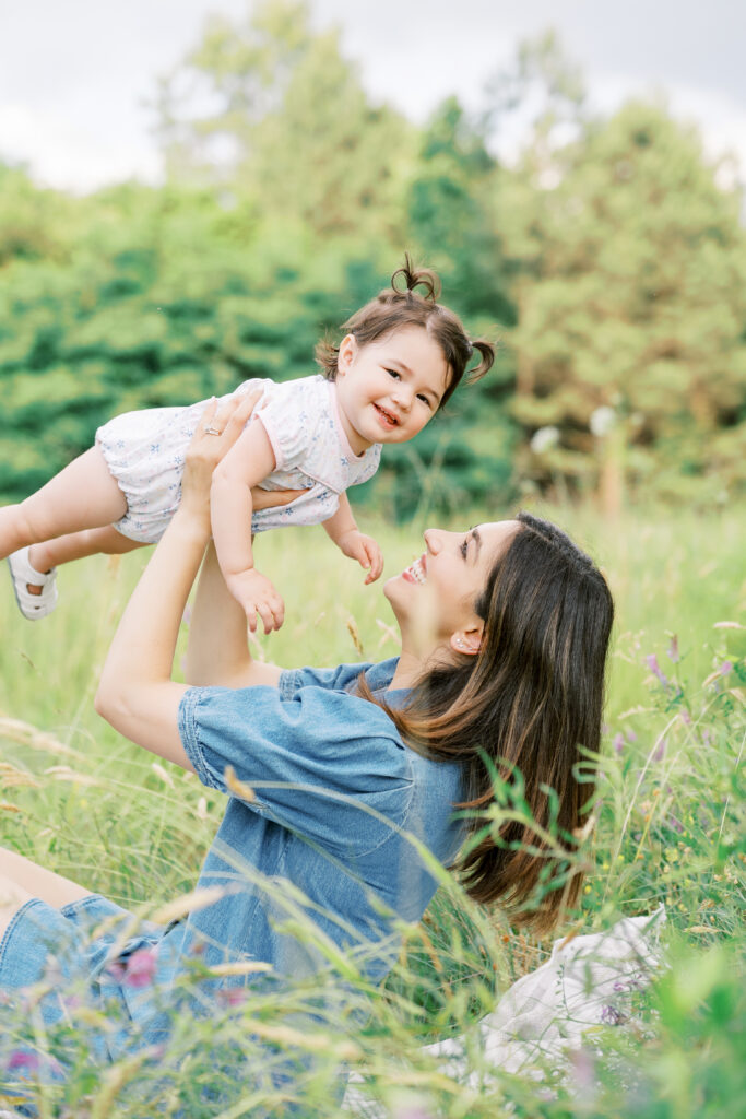 Mom with dark hair wearing a denim dress sits on a blanket in a field with green grass and purple wildflowers and smiles at one year old daughter as she lifts her up in the air.
