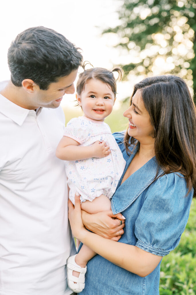 Mom and Dad smile at 1 year old little girl during photos with Raleigh family photographer at the NC Museum of Art Park